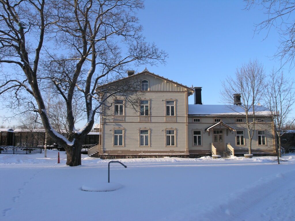 Lumisen maiseman keskellä vaalea, osin kaksikerroksinen rakennus.