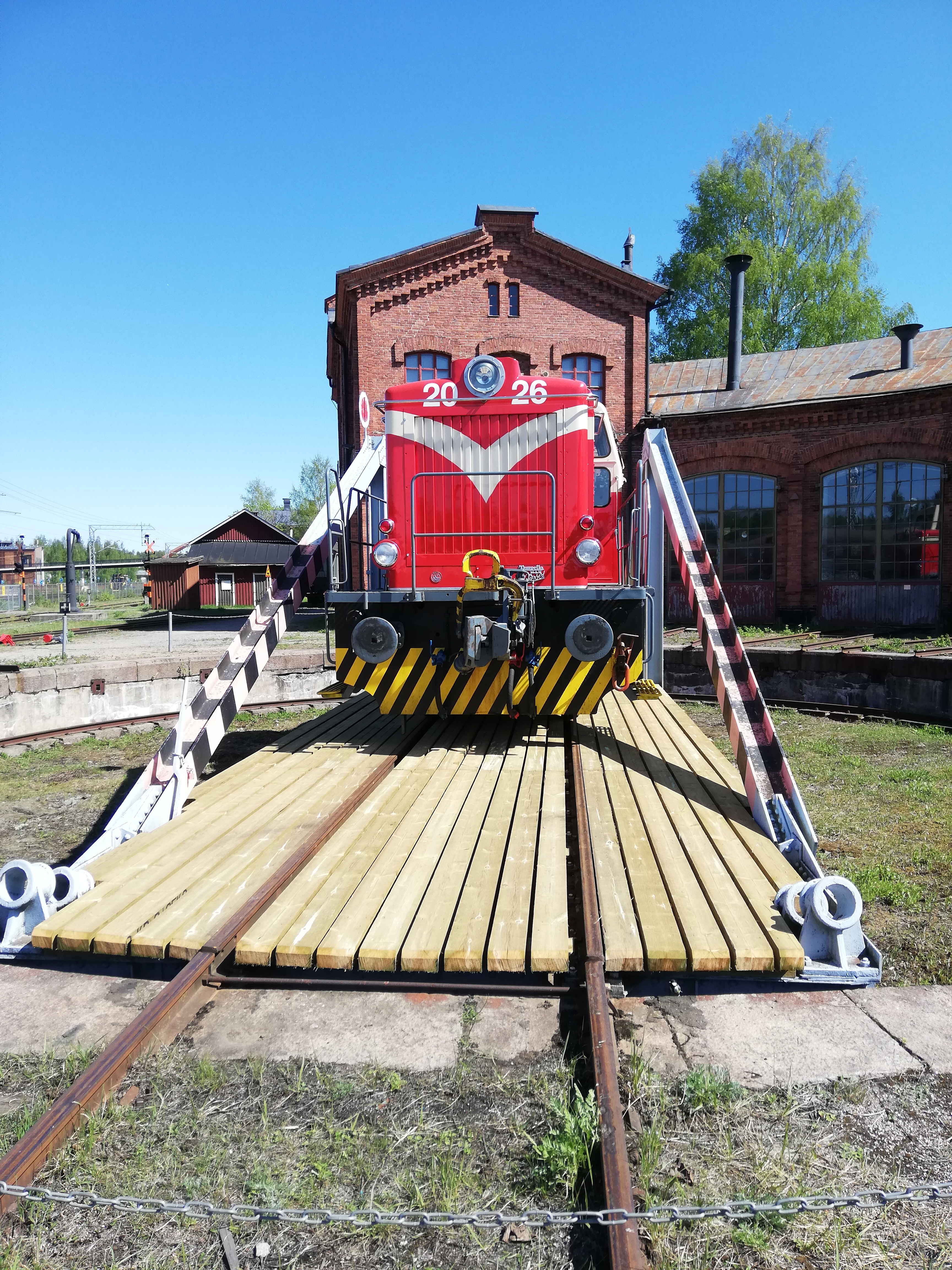 Punainen dieselveturi kääntöpöydällä, taustalla punatiilinen veturitalli.