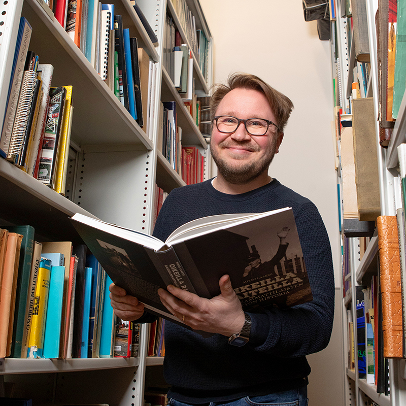 Henkilö lukee kirjaa ja hymyilee kirjahyllyjen välissä.
