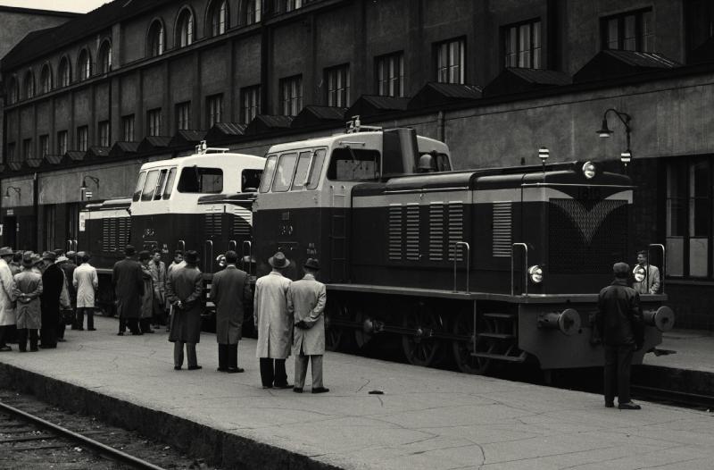 Kaksi veturia rautatieasemalla, vieressä ihmisiä laiturilla.