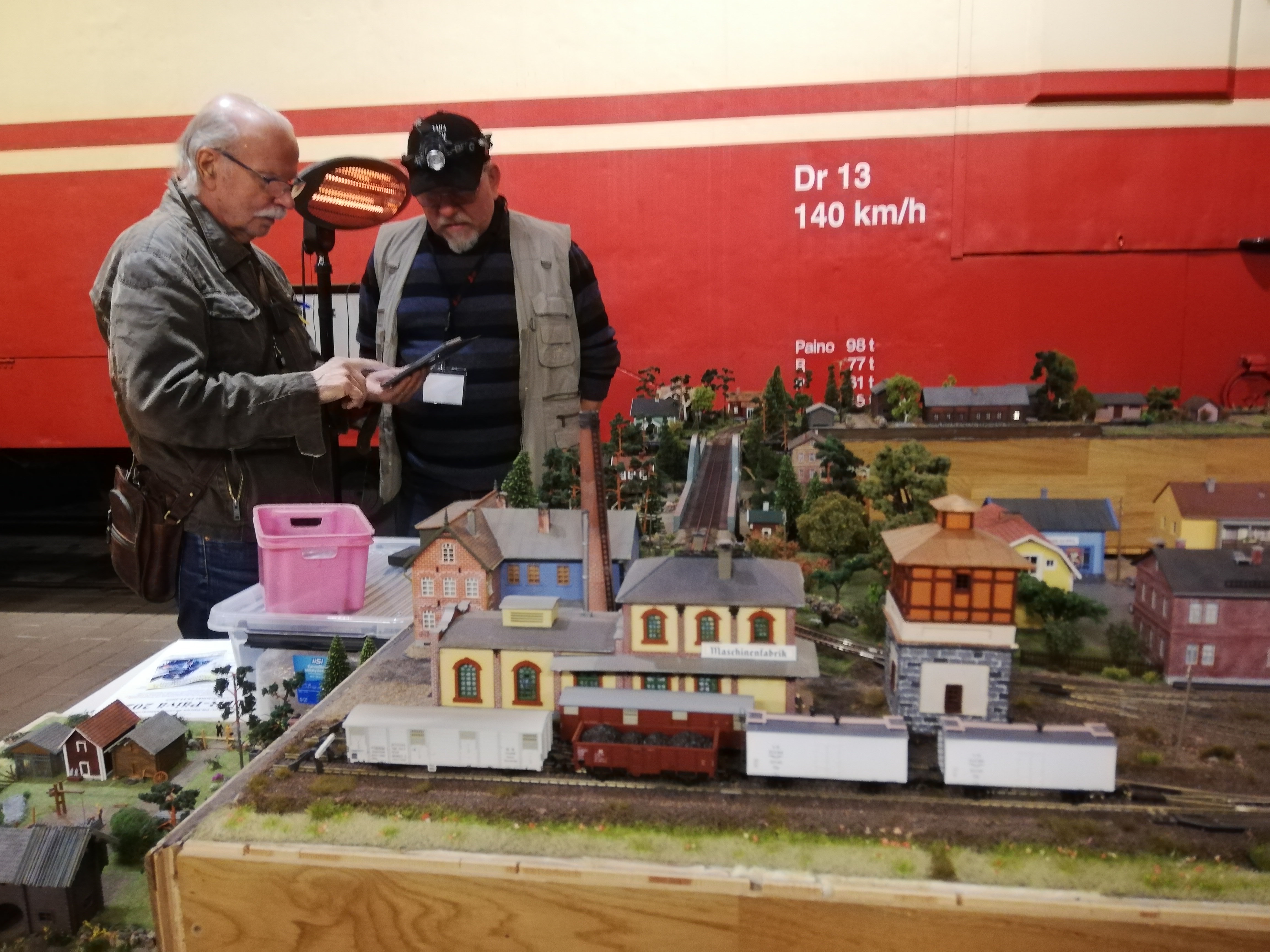 Edessä pöydällä pienoisrautatie, pöydän takana kaksi miestä keskustelee.