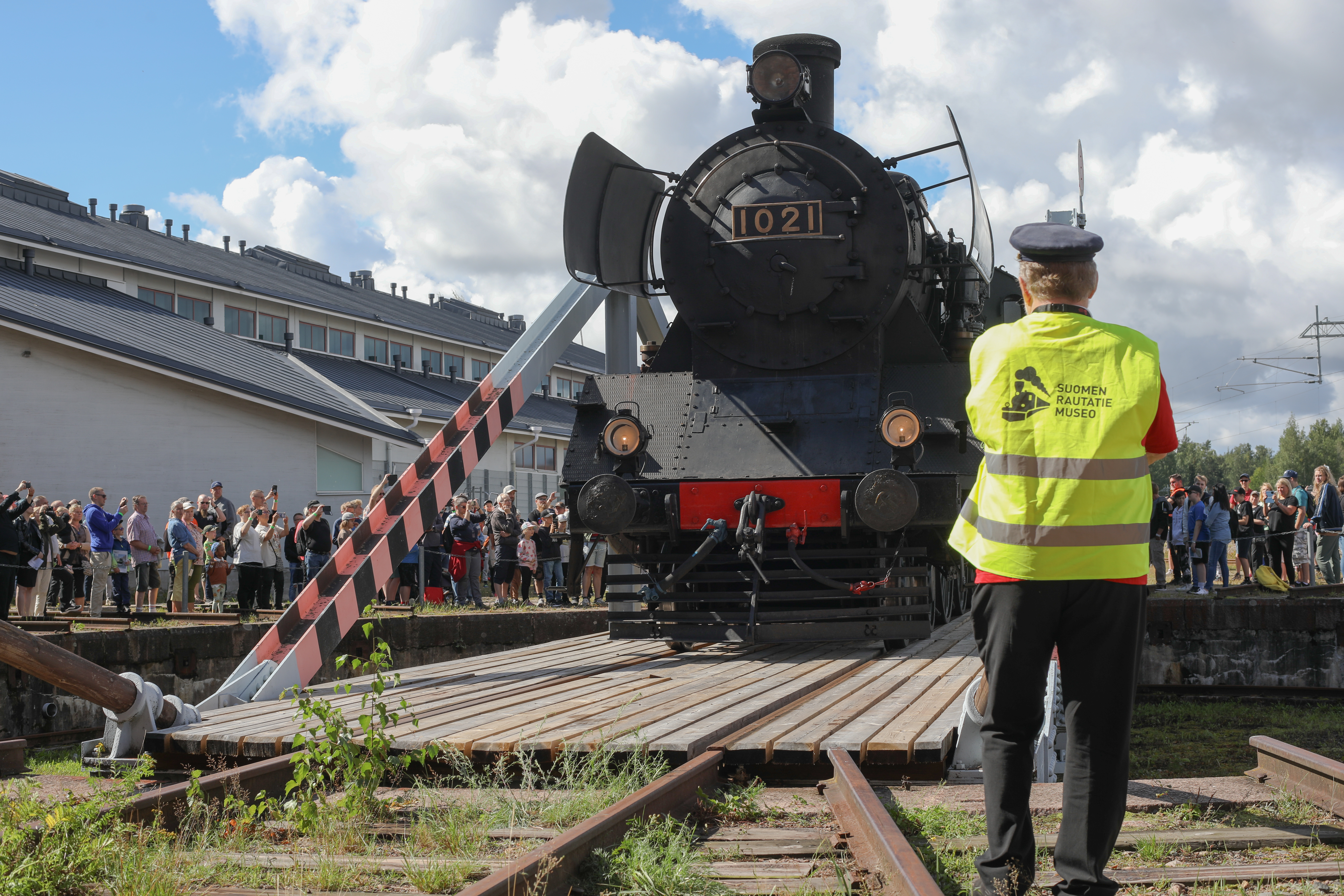 Henkilö selin kameraan, etualalla höyryveturi, taustalla katsojia.